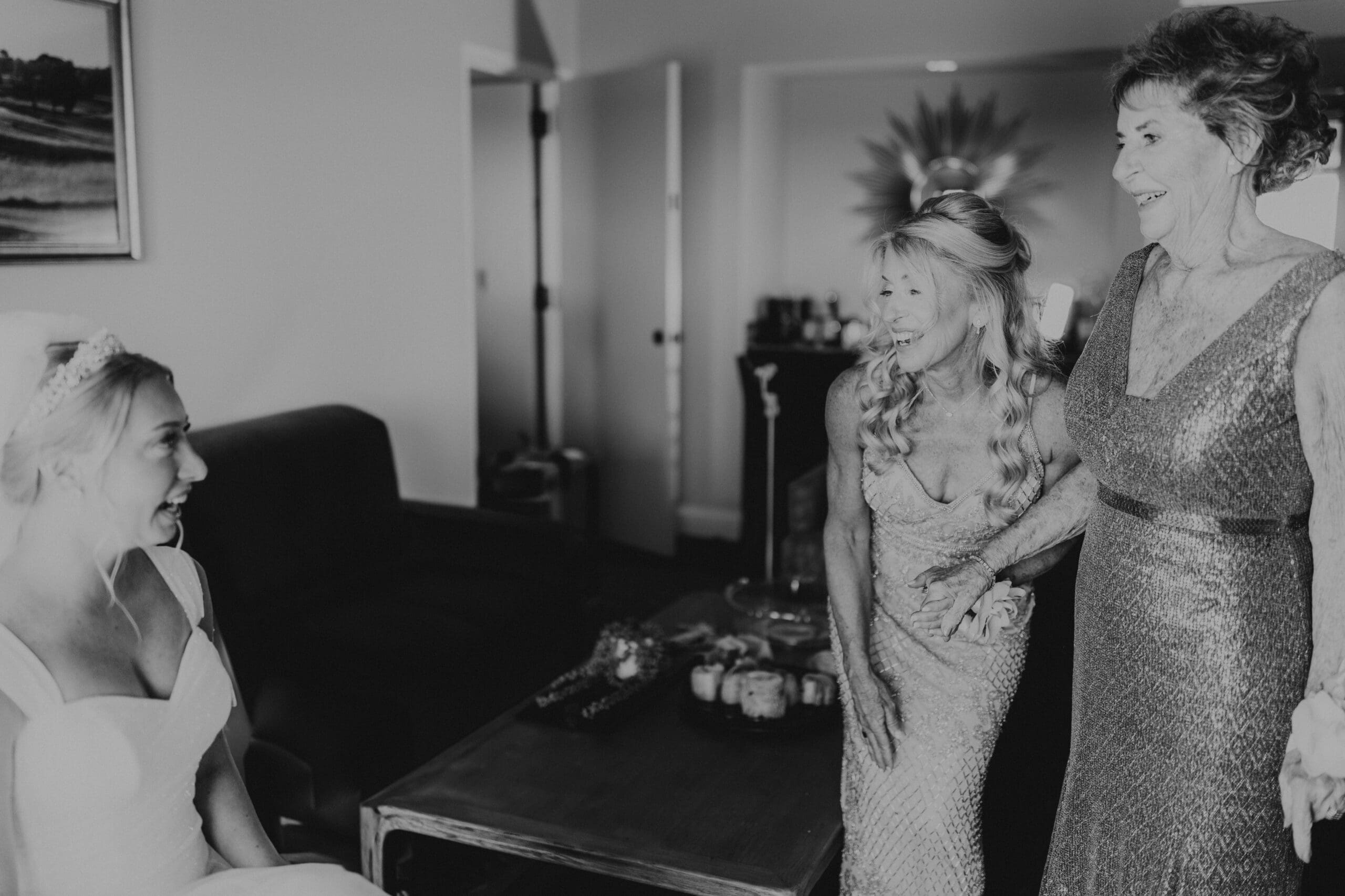 A black and white image of a mother in a long sparkle gown holding onto grandmother's arm, both wearing a corsage and both women beaming at bride who has a giant, open-mouth smile, while sitting on a chair in her wedding gown, with a crown and veil draped behind her head.