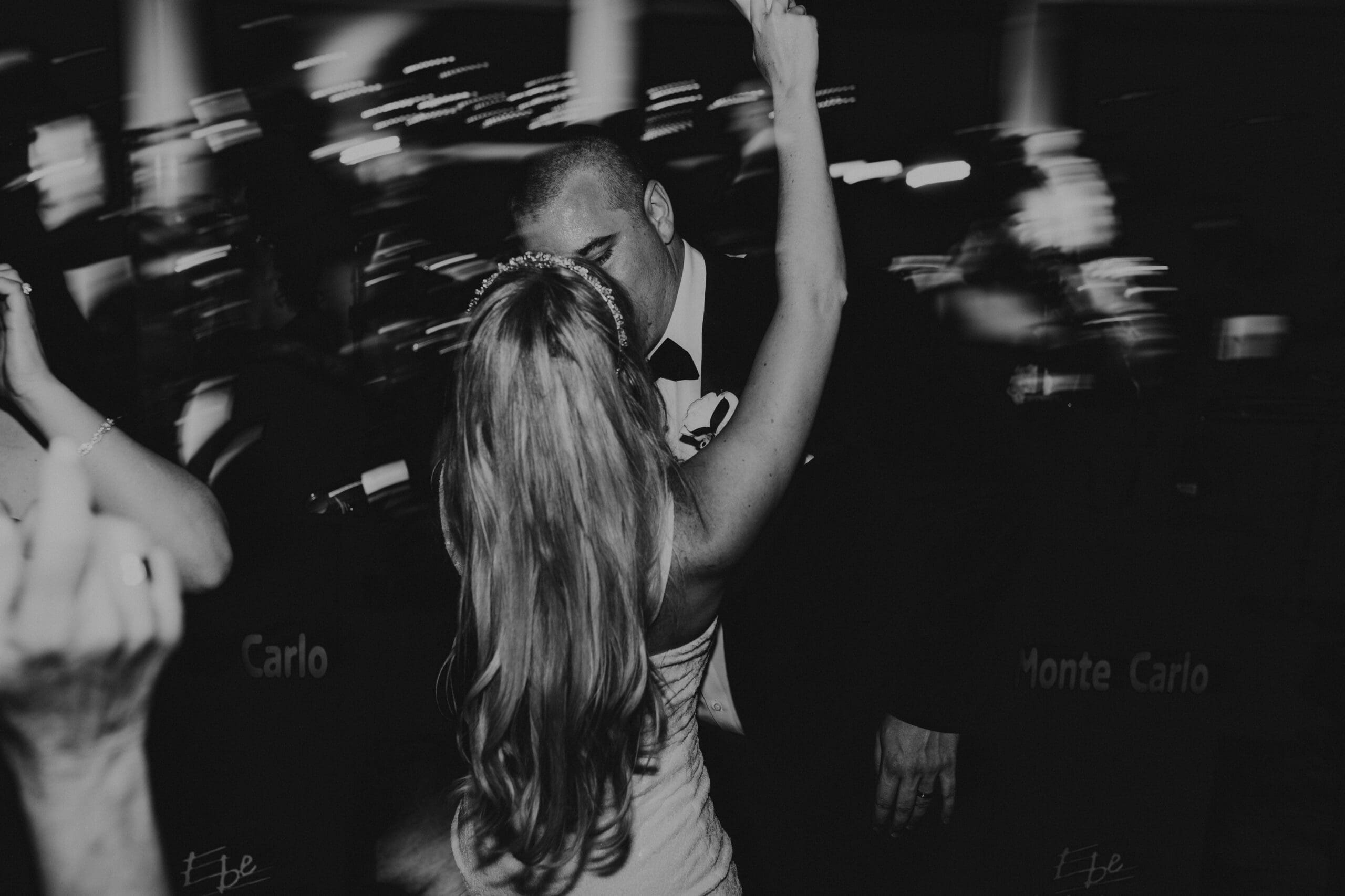 black and white wedding photo of the back of a blonde woman's head, her arm in the air, kissing a man in a black tuxedo, with artistic light drag movement and people dancing all around them.
