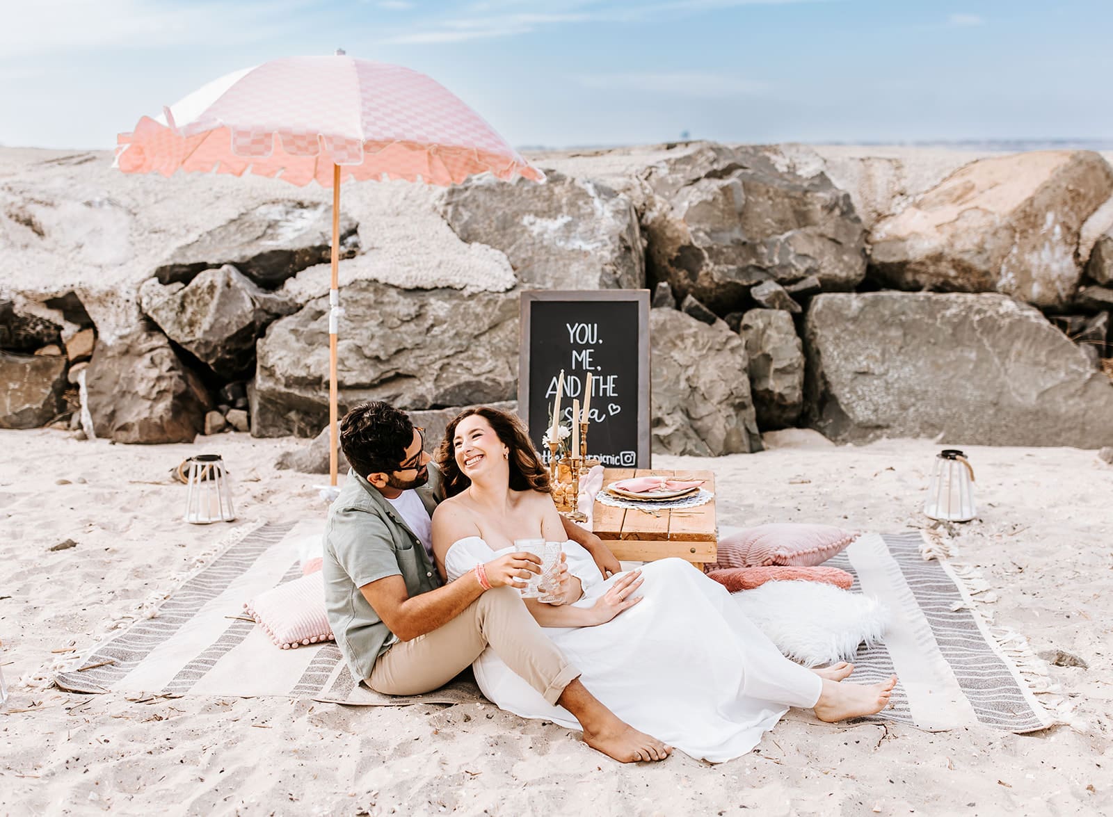 A gorgeous beach sky and giant rocks sit in front of a custom picnic set-up, with man in linen green shirt sitting on a stripped rug with woman in a white flowy dress sits between his legs, both clinking their blush cups together for a cheers.
