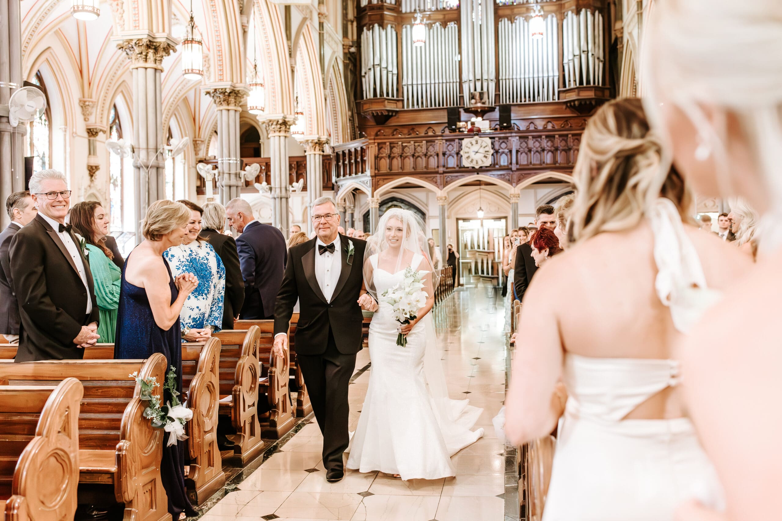 step-father in black tuxedo with white boutonniere walks blushing bride down the aisle as friends and family beam at the sight, inside of historic Philadelpiha church St. John the Baptist.