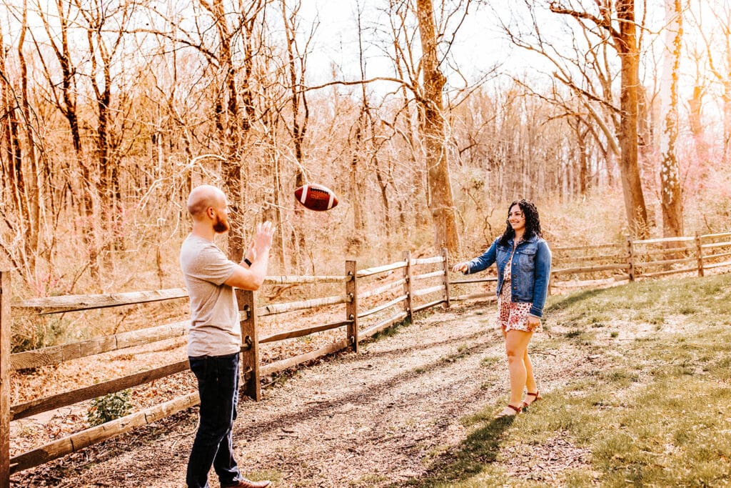 Bald man in tan shirt throwing football to woman in a blue jean jacket, both in front of a wooden fence and trees with no leaves.
