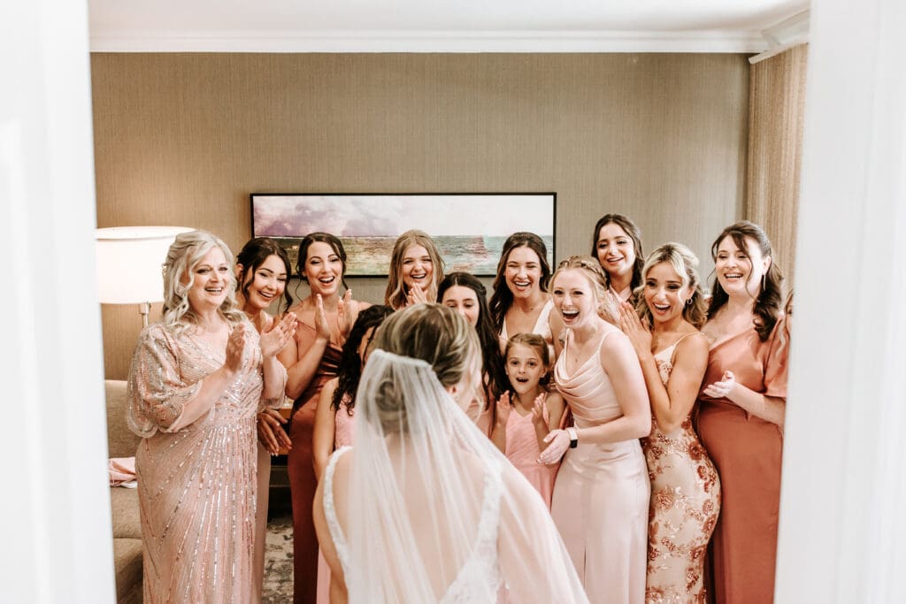 Group of bridesmaids in an array of blush dresses are shocked and brought to tears by the vision of the bride on her wedding day.
