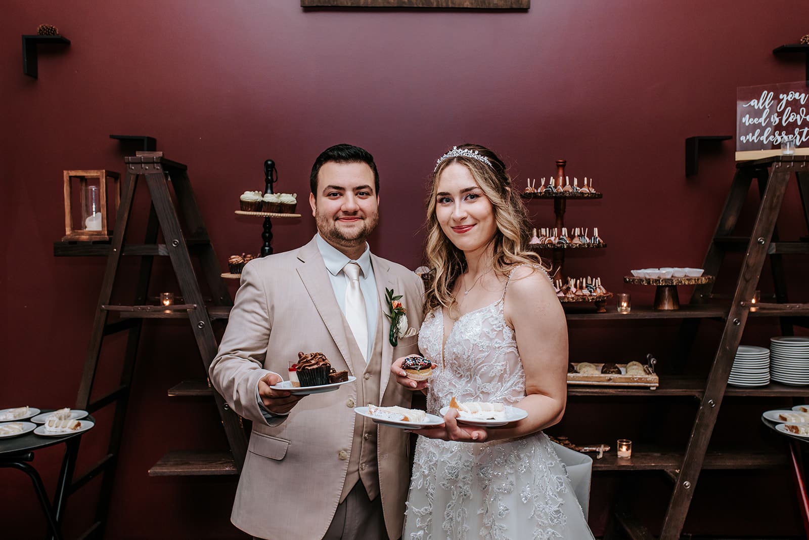 Bride and Groom happily smiling at camera, together holding dessert plates in Running Deer Golf Club's dessert room, surrounded by sweet treats.
