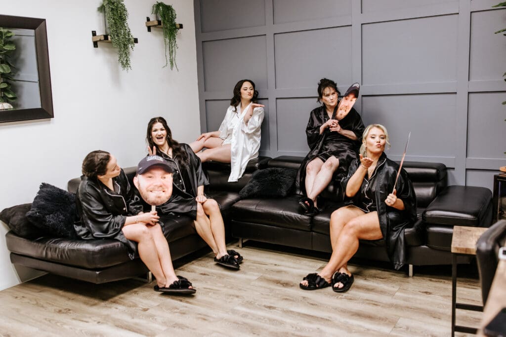 Bride and her bridal party share a fun, playful moment in black satin robes while getting ready. The bride, in a white robe, blows a kiss while the bridesmaids hold oversized cutouts of the groom’s face and laugh together in a modern lounge setting.