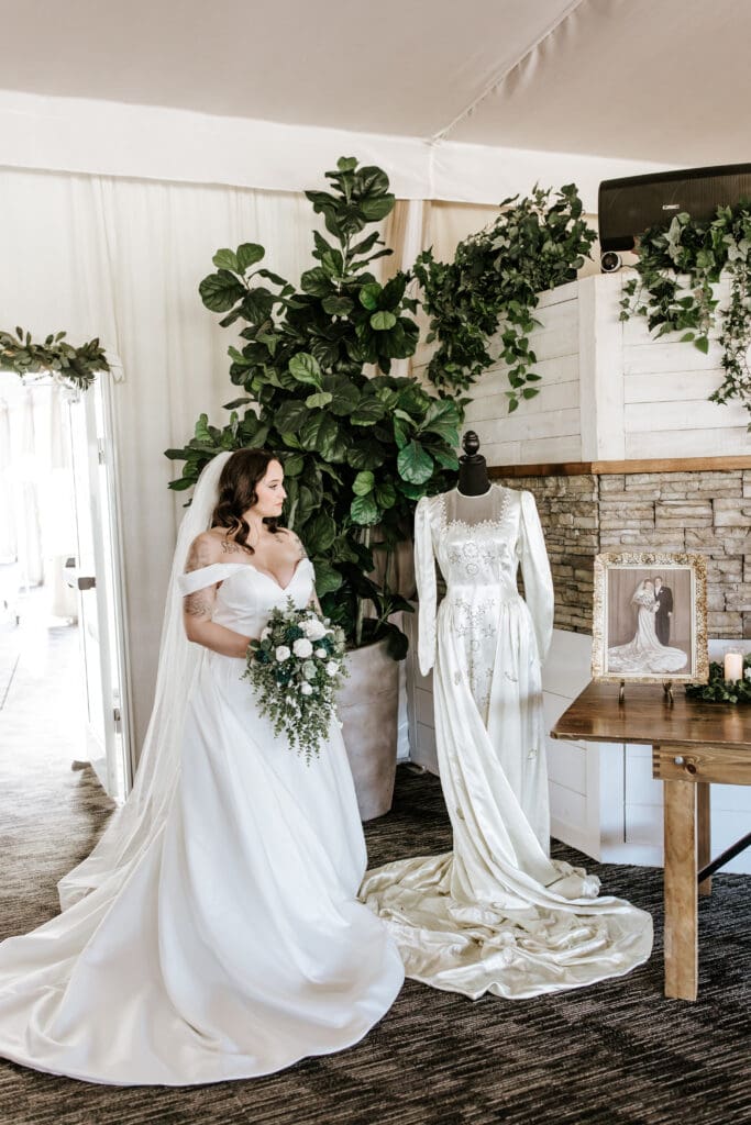 Bride in a modern off-the-shoulder gown stands beside a vintage wedding dress display, holding a white and green bouquet. The scene is elegantly decorated with lush greenery, a framed photo of a past wedding, and a candle on a rustic wooden table, evoking a sense of generational love and legacy.
