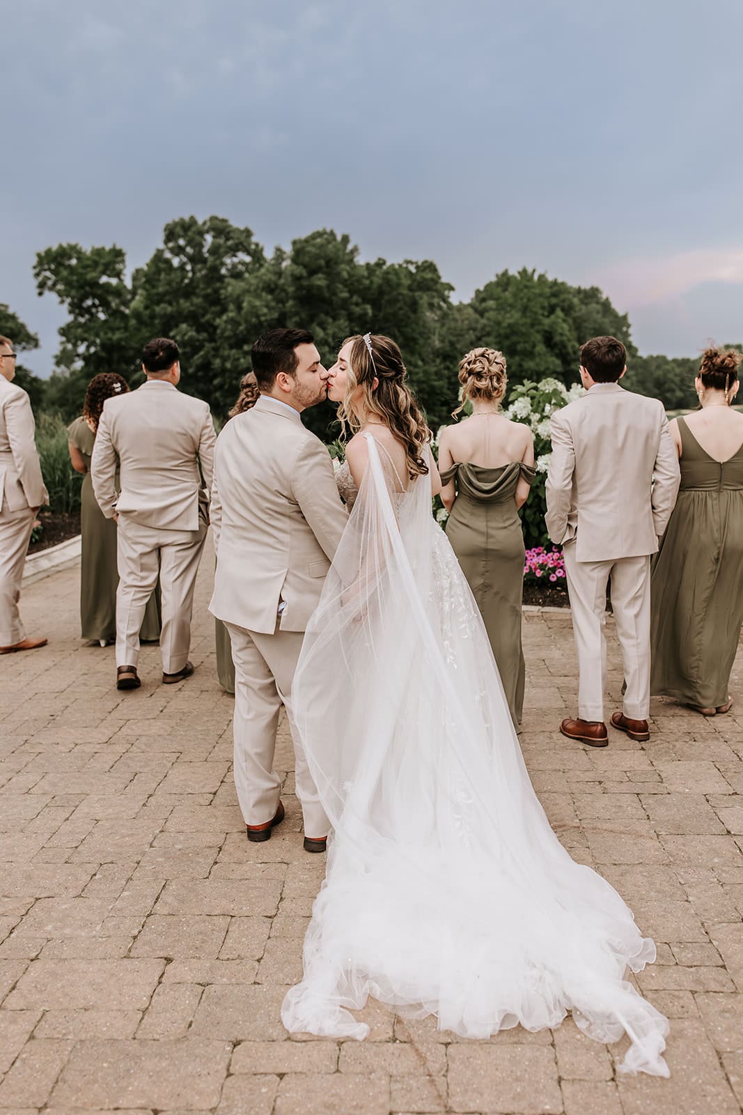 Running Deer Golf Club Weddings Kayla Aspen Photography Groom in tan suit kissing bride with cape draped over her shoulder in her wedding dress with their bridal party walking in front of them over the hills at Running Deer Golf Club.