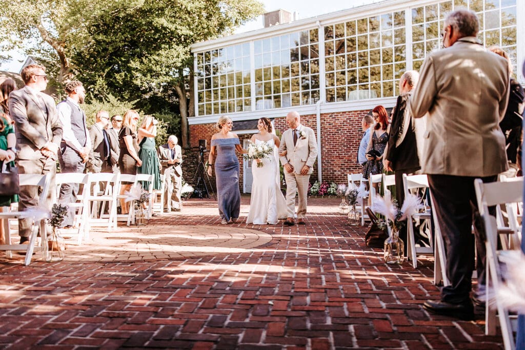 Mom in off-the-shoulder blue gown and corsage, joyfully holding onto bride as dad in tan suit is on the opposite side, both holding onto the brides arms, asa they walk down the aisle at Historic Smithville Inn. Wedding guests are standing and smiling from both sides of the aisle. 