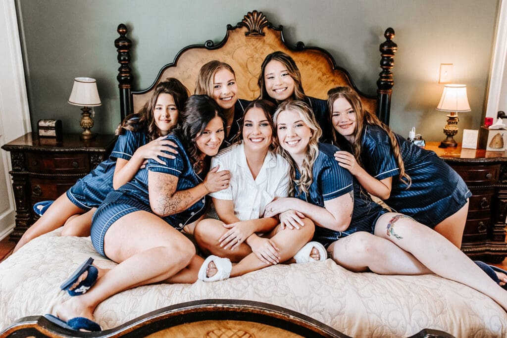 Group of bridesmaids smiling in navy satin striped pajamas sitting on Historic Smithville Bed and Breakfast's bed, with arms together holding the bride, who is sitting in the middle with a short-sleeve white button up pajamas set and white fluffy slippers. 