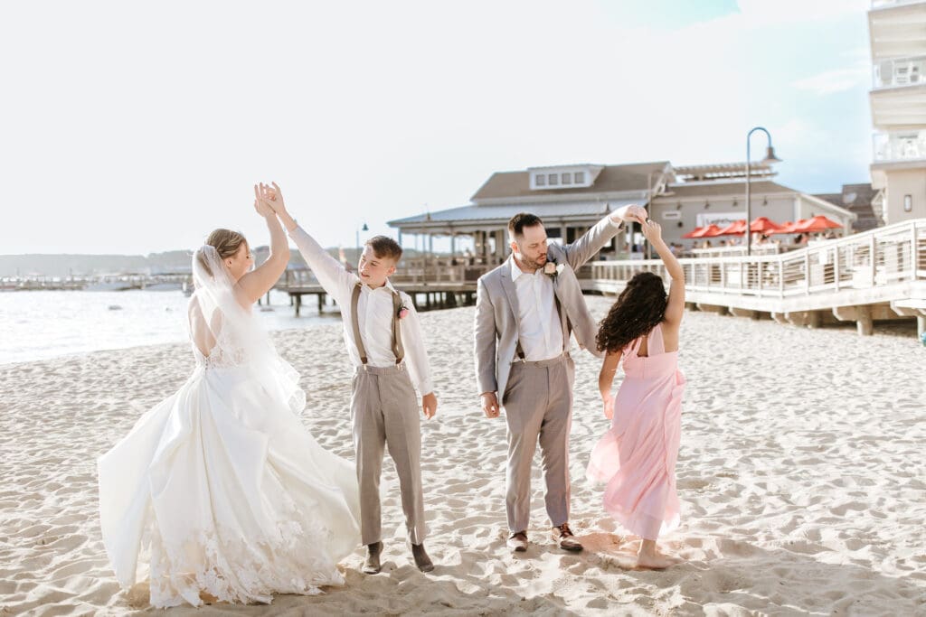 Boy in suit white shirt with suspenders is twirling his new step-mother, who is dressed in a beautiful white lace wedding gown and veil. Next to them is new step dad wearing a gray suit, twirling his new step-daughter, who is wearing a blush dress. The four of them are on the sand, in front of Lighthouse Cover Event Center in Dewey Beach, DE. 