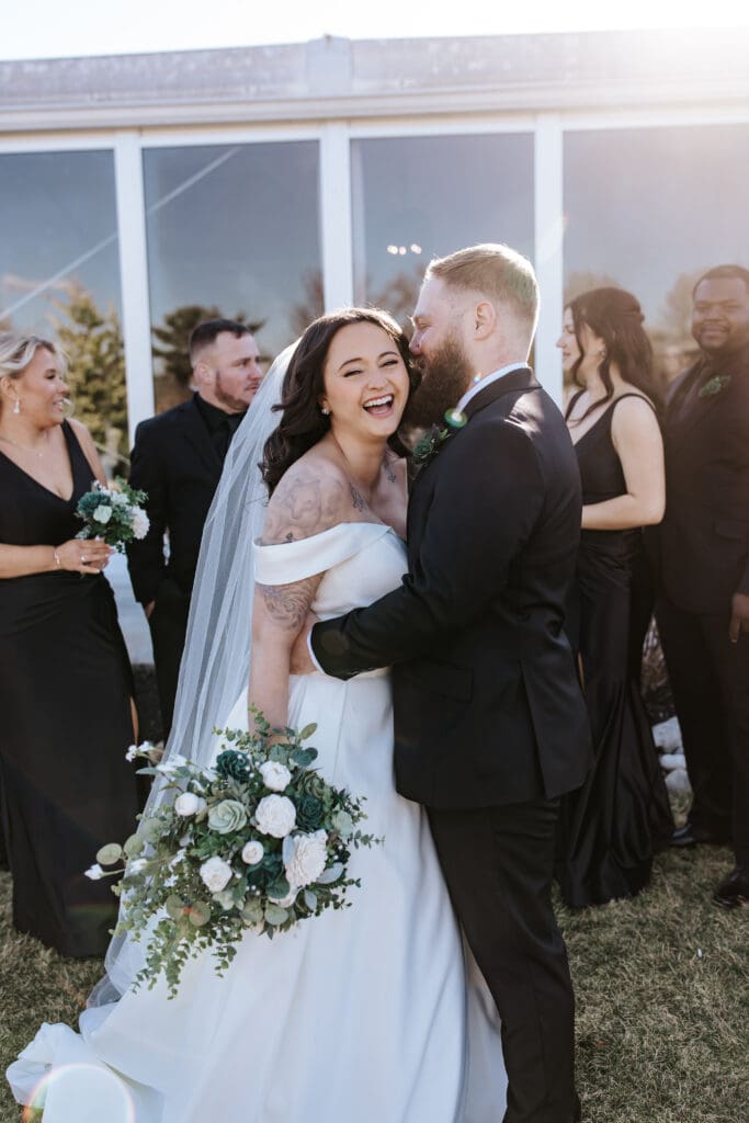 Bride and groom share a happy moment surrounded by their wedding party. The bride laughs as the groom kisses her cheek, holding a bouquet of white and green florals, with sunlight softly filtering through the scene.