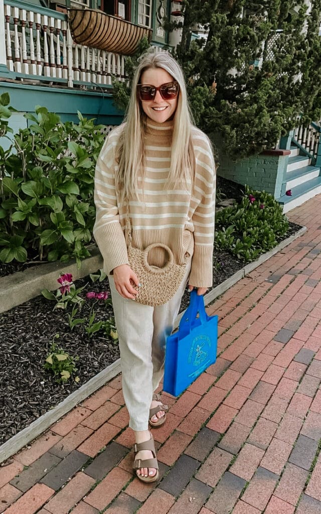 Woman in a neutral striped sweater and linen pants holding a bright blue shopping bag while walking along the Washington Street Mall in Cape May, New Jersey. Casual spring outfit with Birkenstock sandals and a woven crossbody bag.