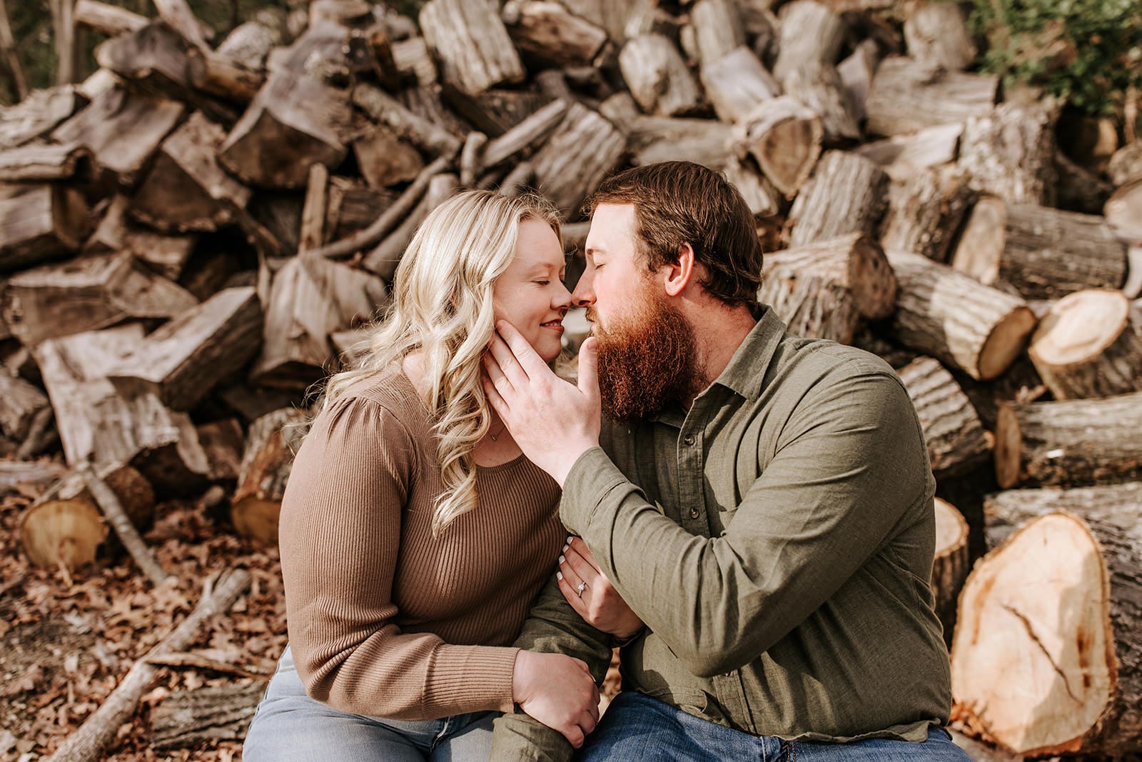 Engaged couple snuggles in front of a stacked woodpile during their South Jersey engagement session, sharing a tender moment on the site of their future home.