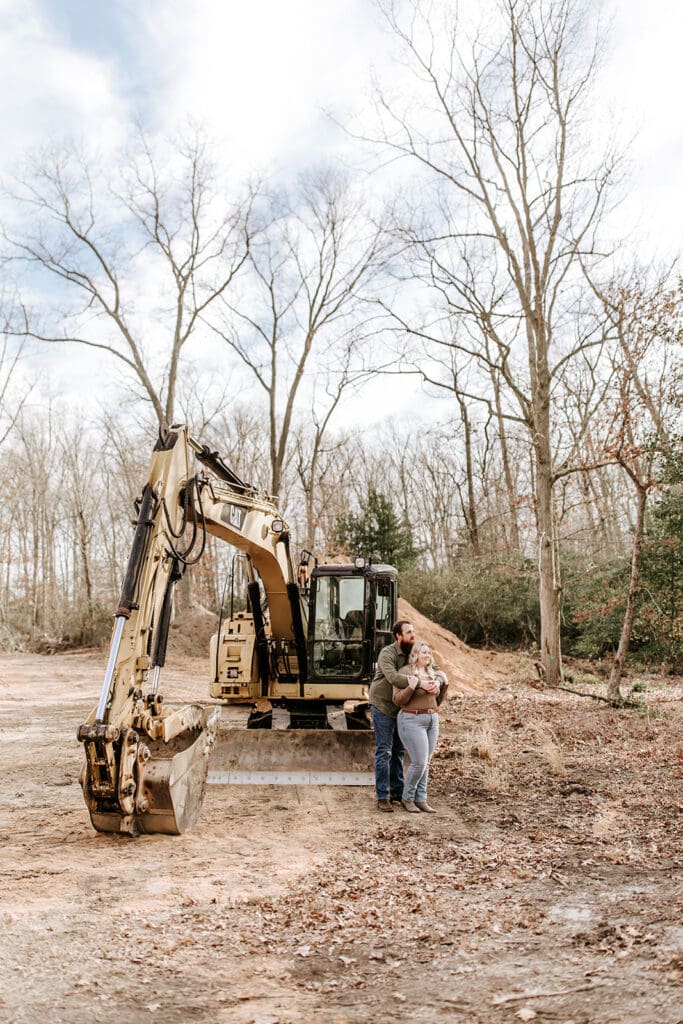Couple embracing in front of construction equipment on their land during their engagement session in South Jersey, celebrating the future site of their new home.