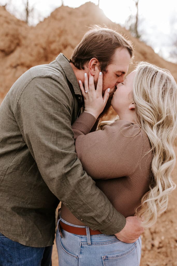 Close-up of a couple sharing a kiss, the bride-to-be’s engagement ring visible as sunlight hits the sand pile behind them during their South Jersey engagement session.