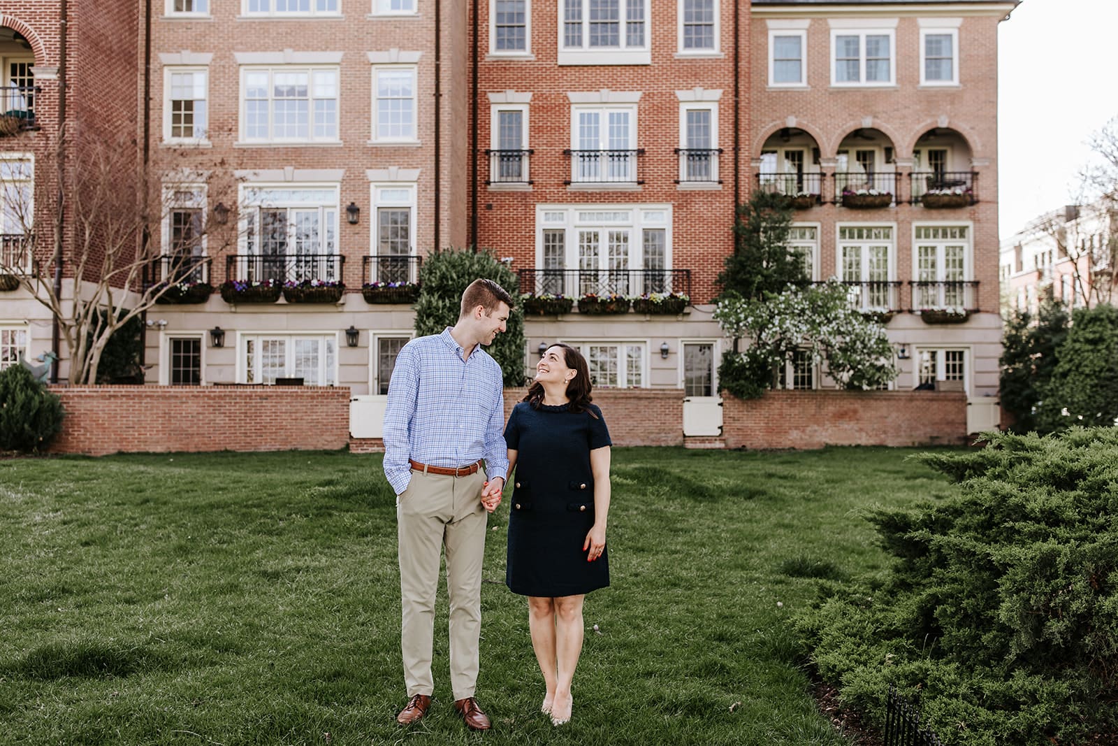 Couple holding hands and smiling in front of charming brick townhomes in Old Town Alexandria, standing on a lush green lawn during their spring engagement session.