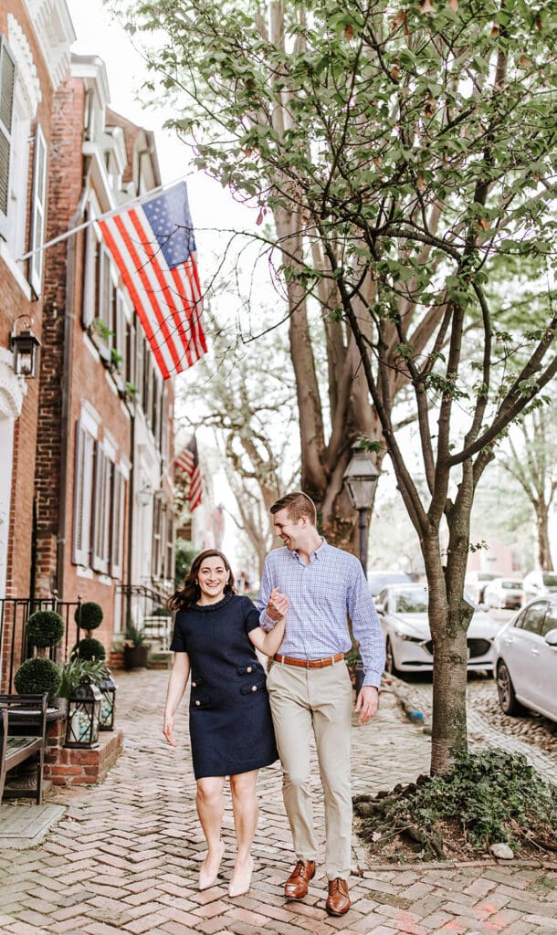 Couple walking hand-in-hand down the historic cobblestone streets of Old Town Alexandria during their engagement session, with American flags and spring trees lining the charming red-brick townhomes.