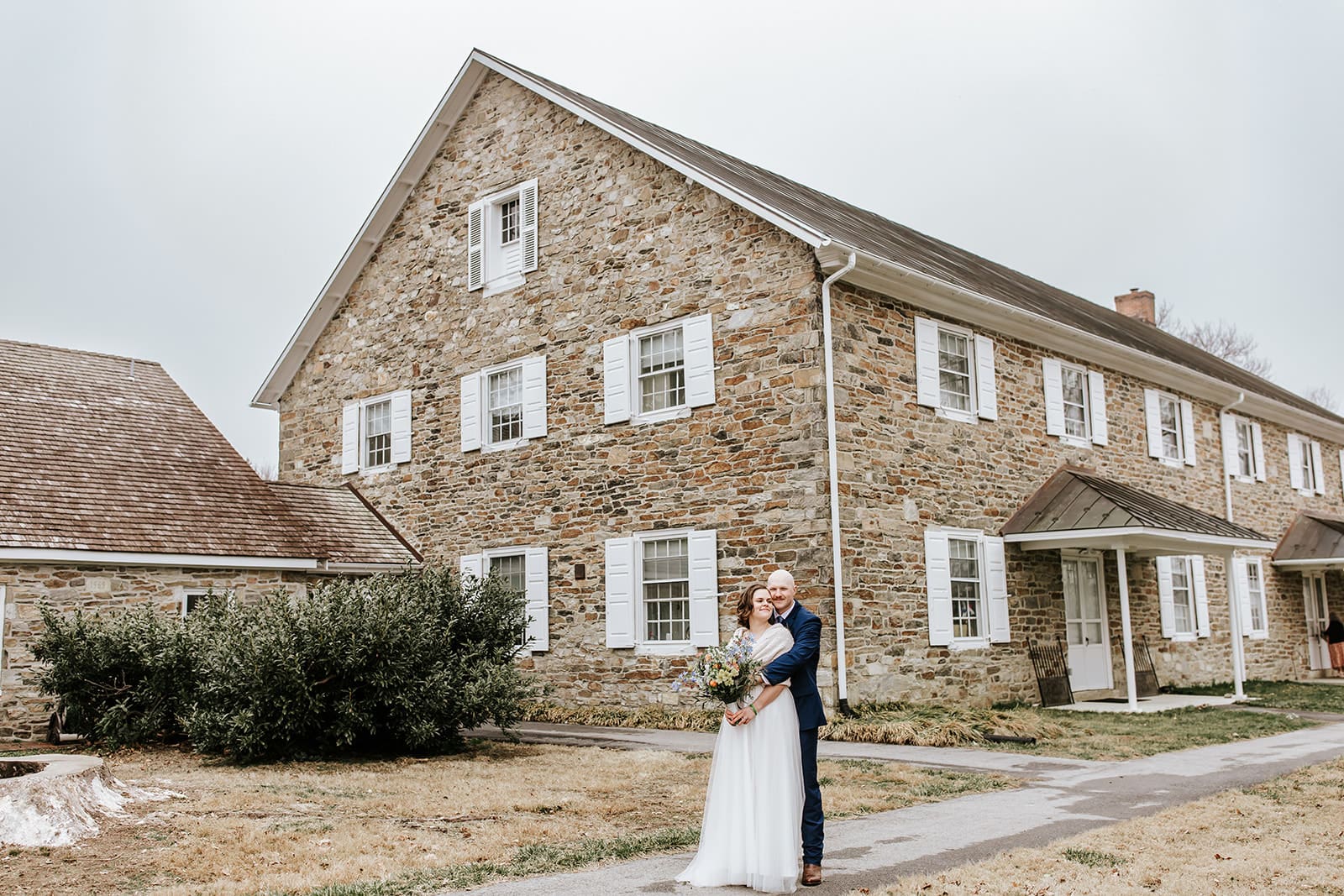 Bride and groom embrace in front of the historic stone London Grove Quaker Meeting House in Kennett Square, Pennsylvania, during their intimate Chester County wedding day.