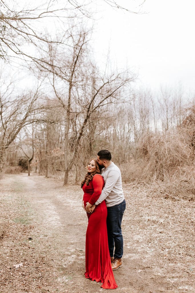 Expecting couple embracing during a winter maternity session at Riverwinds in South Jersey. The mom-to-be wears a long red gown while her partner holds her from behind on a peaceful wooded trail.