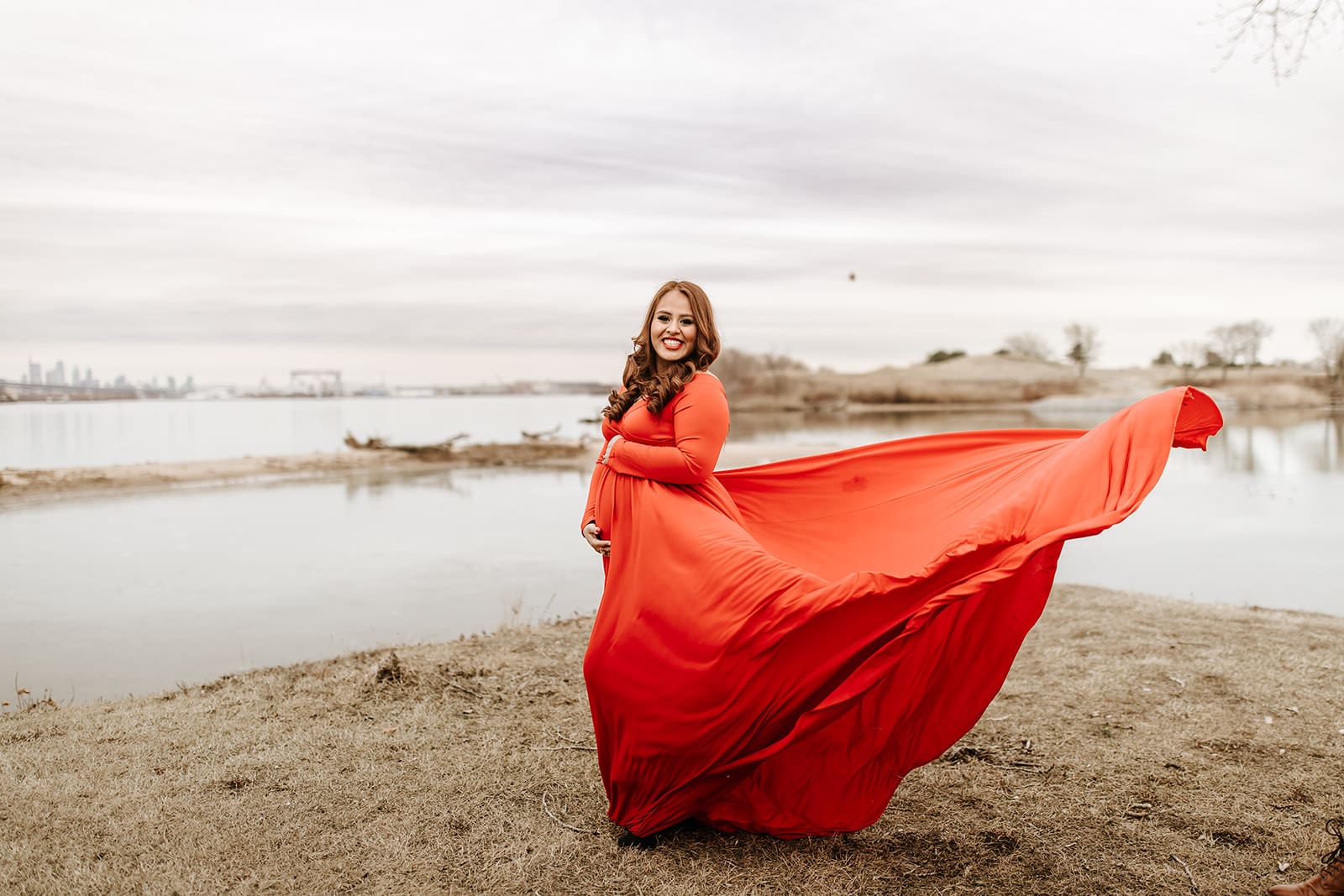 Maternity photo of a glowing mom-to-be in a flowing red gown by the water at Riverwinds in South Jersey. The Philadelphia skyline is visible in the distance, adding a dreamy backdrop to this waterfront maternity session.