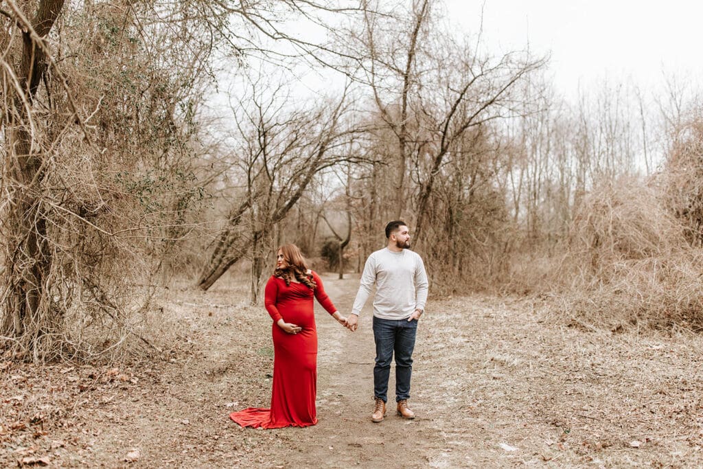 Couple holding hands and standing apart on a wooded path during their Riverwinds maternity session in South Jersey. The mom-to-be wears a red gown, highlighting the anticipation of their baby boy arriving in June.
