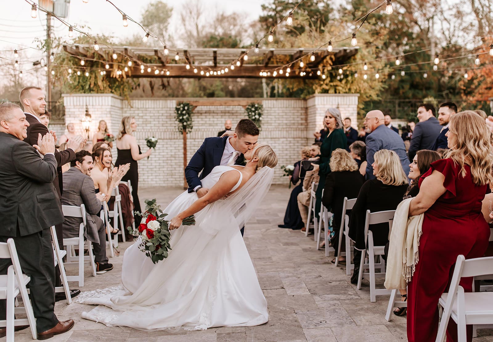 Bride and groom kiss at the end of their outdoor ceremony under twinkle lights at The Grove at Centerton, surrounded by clapping guests and warm fall tones.