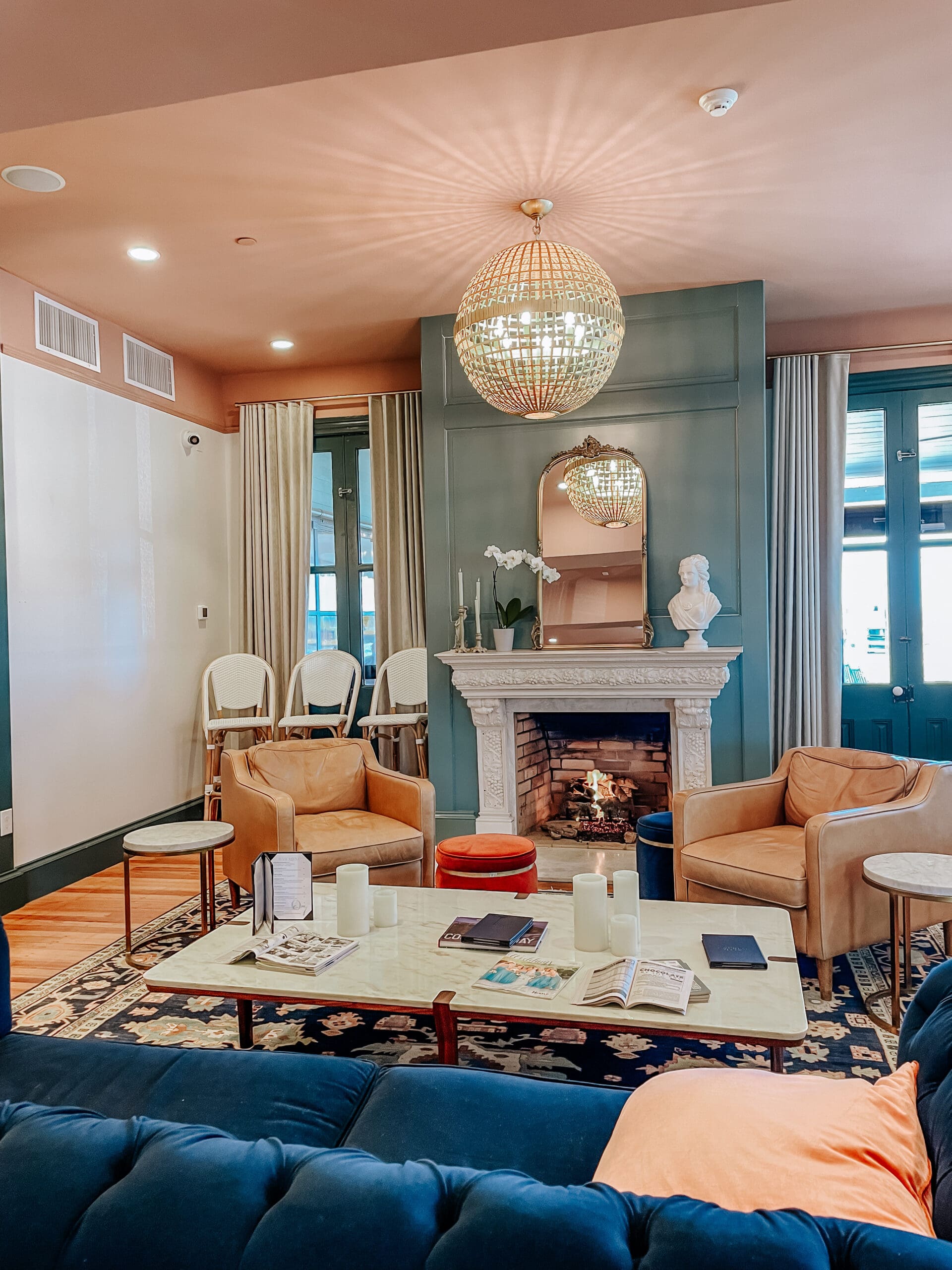 Cozy and elegant sitting room inside The Inn of Cape May, featuring a warm fireplace, velvet and leather seating, a marble coffee table with books and candles, and a statement chandelier reflecting soft light onto the peach-toned ceiling.