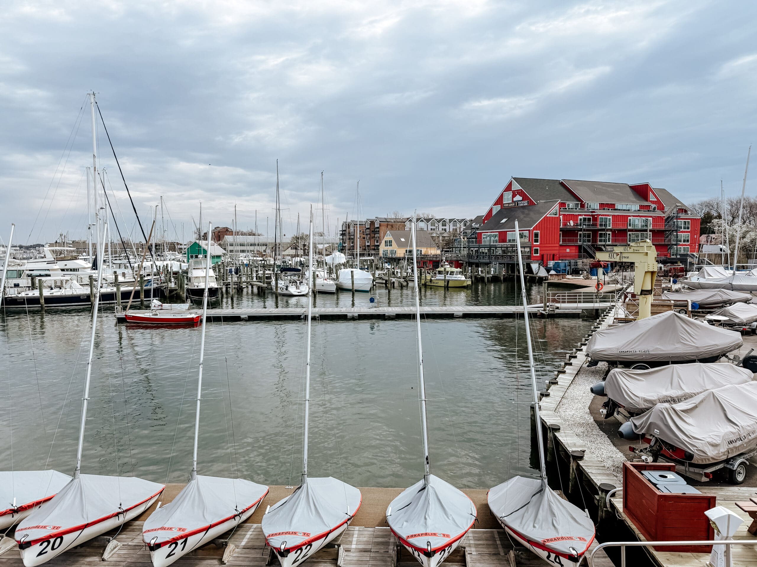 View of a marina in Annapolis, Maryland featuring docked sailboats, covered boats, and the iconic red Annapolis Waterfront Hotel in the background under a cloudy sky.