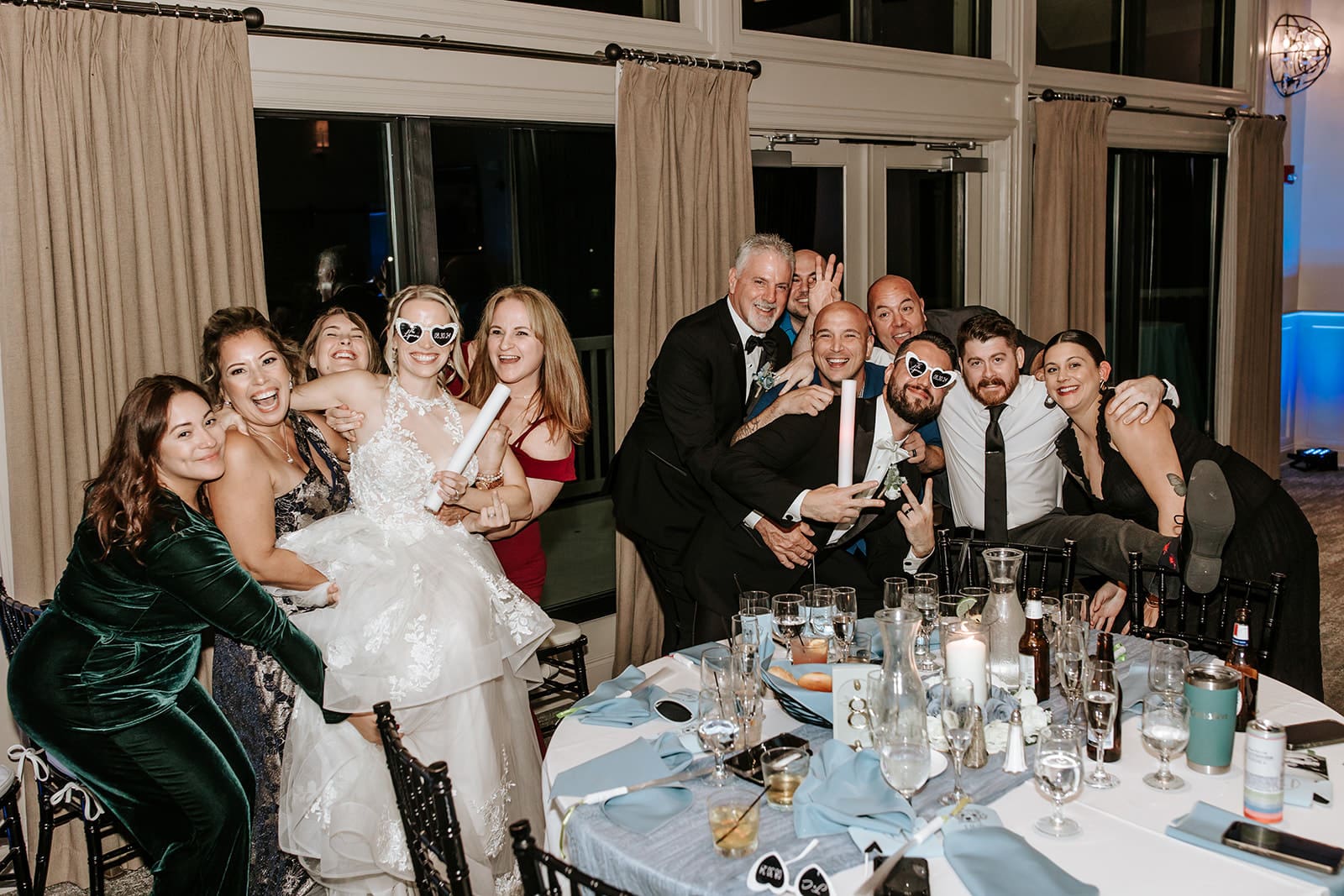Bride wearing heart-shaped sunglasses celebrating with wedding guests around a reception table during fun table photos at a New Jersey wedding.