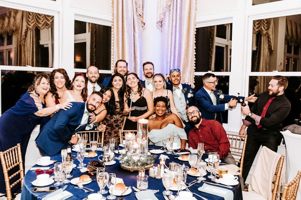 Wedding guests posing with the bride and groom during fun table photos at a New Jersey wedding reception with navy blue table decor and candle centerpieces.