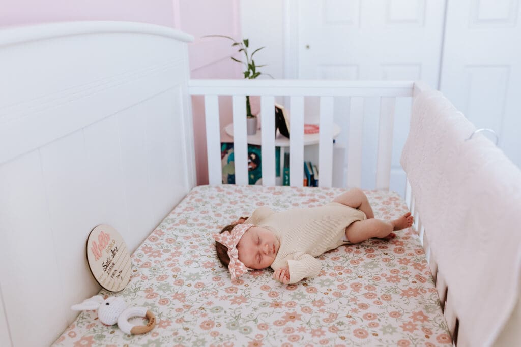Newborn baby girl Sadie sleeping peacefully in a white crib with a floral sheet, wearing a neutral onesie and pink bow headband. A personalized wooden name sign rests beside her. Captured during an in-home newborn session in Pottstown, PA by Chester County photographer Kayla Aspen.