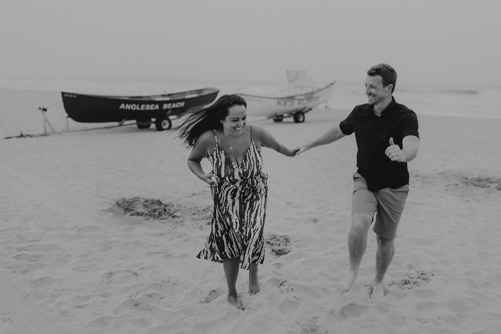 Black and white photo of an engaged couple laughing and running barefoot on the beach in North Wildwood, New Jersey. Two classic lifeguard boats marked “Anglesea Beach” and “North Wildwood” are visible in the background.