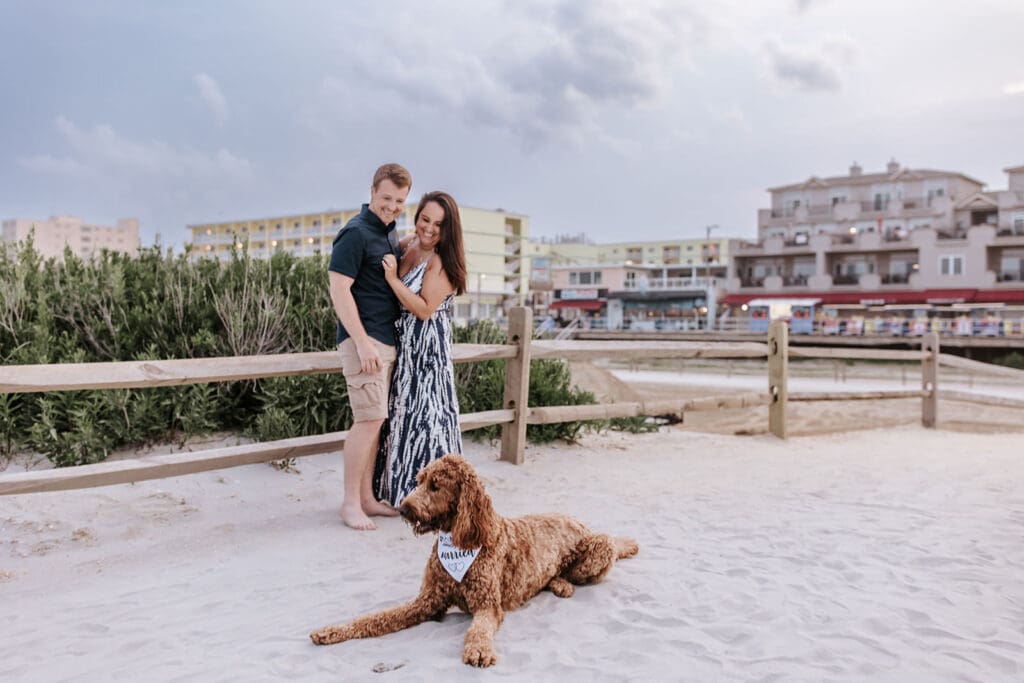 Engaged couple laughing together on the beach in North Wildwood, New Jersey with their goldendoodle, Clifford, laying in the sand wearing a “We’re Getting Married” bandana. Stormy skies and Morey's Piers in the background.