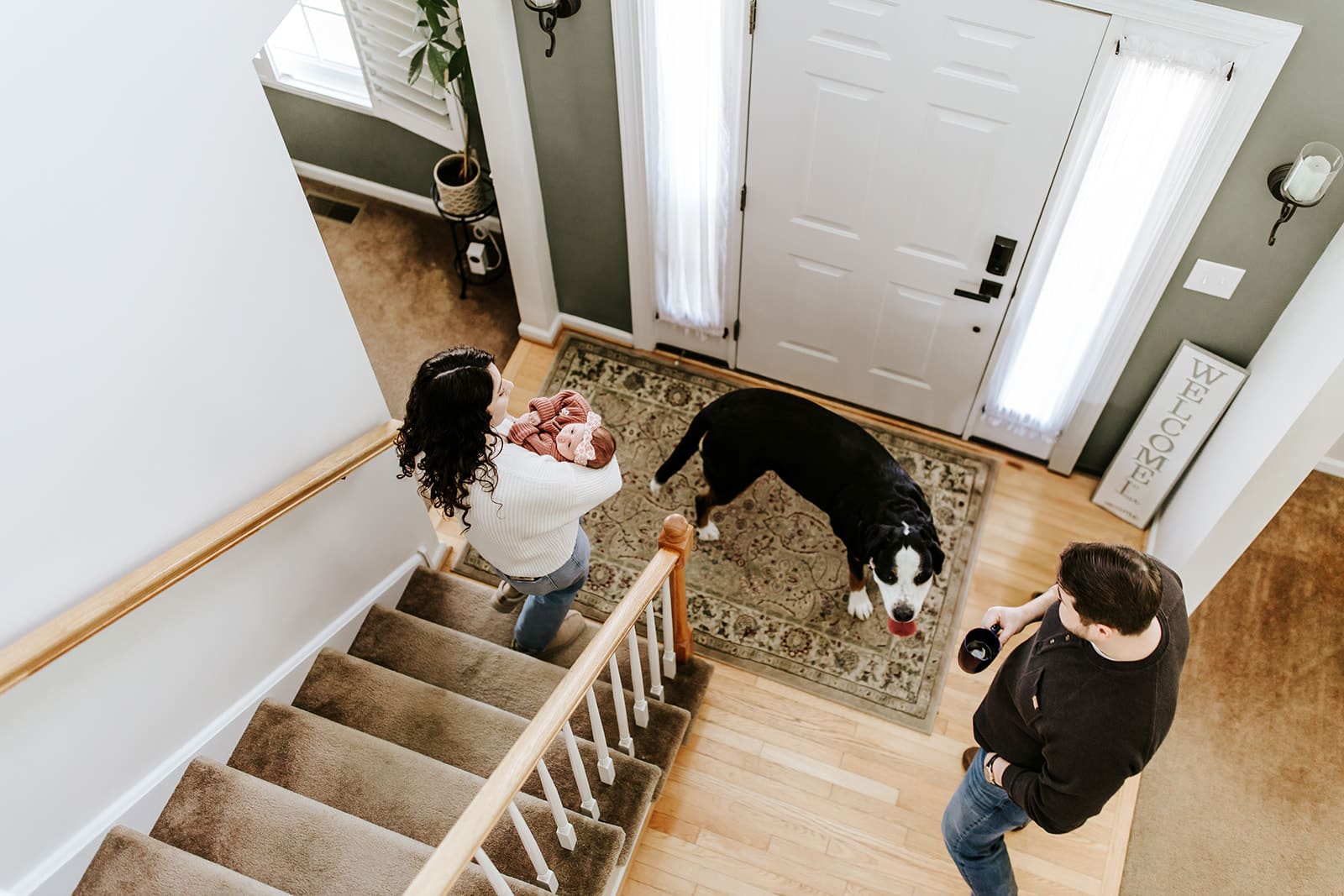 Overhead view of a mother carrying her newborn baby girl Sadie up the stairs during an in-home newborn session in Pottstown, PA, with dad standing nearby and their large black and white dog Dunkin waiting at the front door. Captured by Chester County newborn photographer Kayla Aspen.