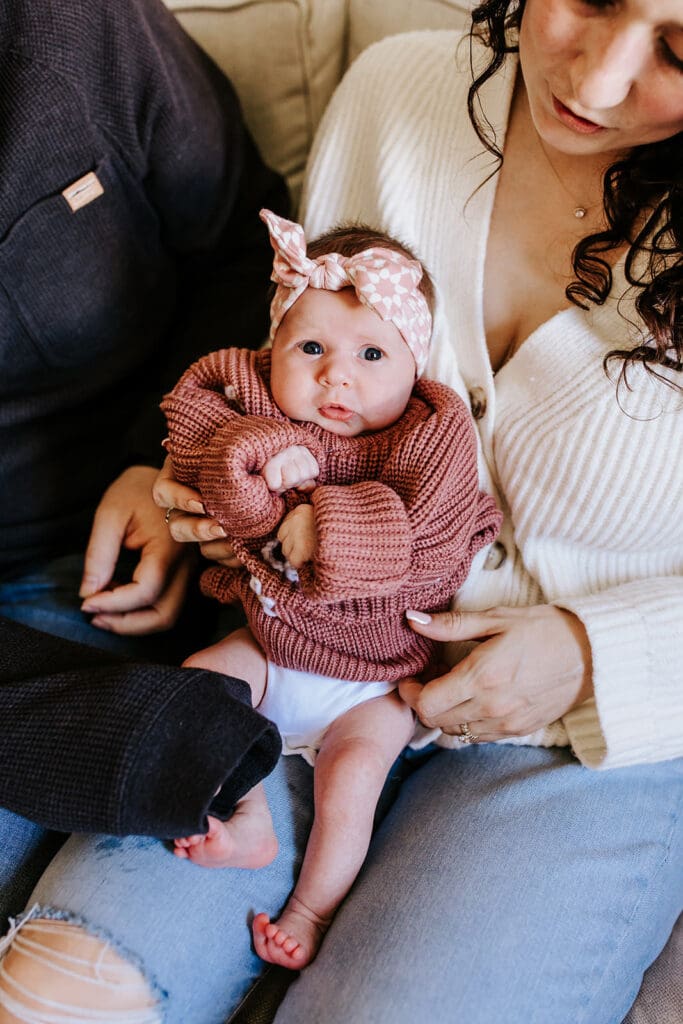 Newborn baby girl Sadie in a cozy rose knit sweater and pink headband, held by her parents during an in-home newborn session in Pottstown, PA. Captured by Chester County newborn photographer Kayla Aspen.