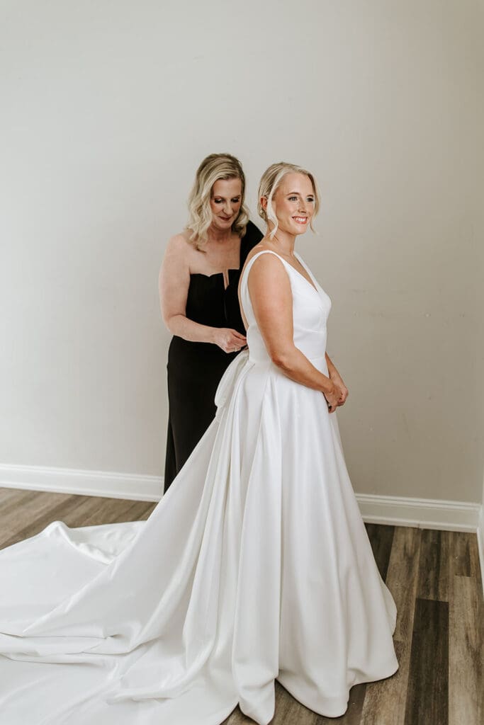 Bride getting ready at The Grove at Centerton as her mom helps zip her classic satin wedding gown.