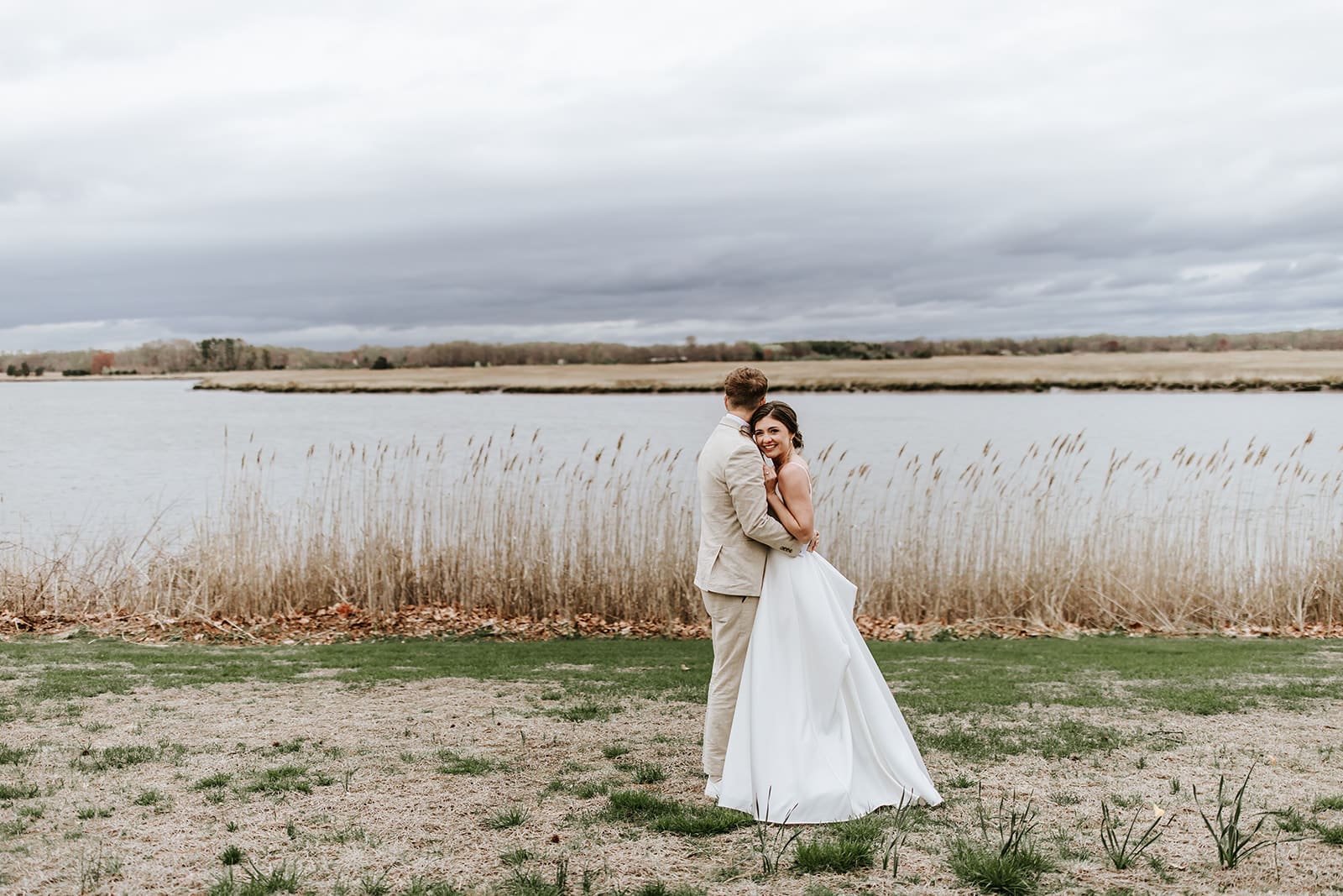 Bride and groom sharing a quiet moment by the river at Eagle Manor in Bridgeton, New Jersey, with tall grasses and moody skies in the background.