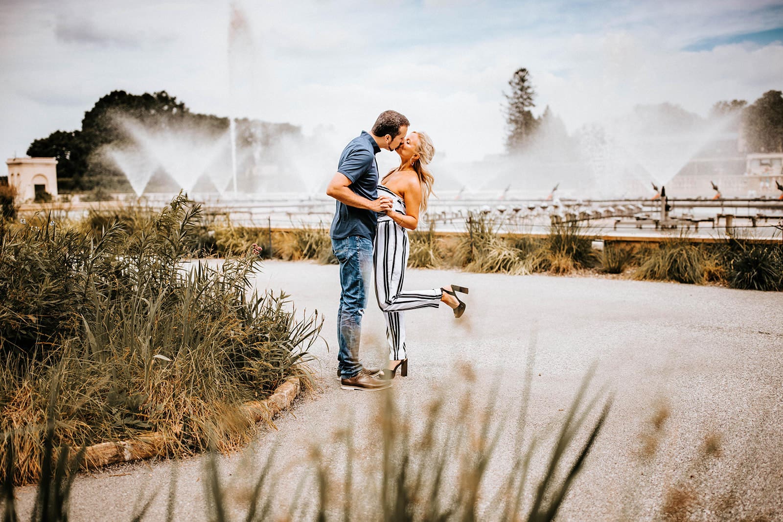 Couple shares a joyful kiss in front of the fountains at Longwood Gardens during a summer engagement session. The woman wears striped pants and heels, lifting one foot playfully as they hold hands. A light-hearted and elegant moment perfect for intentional couples seeking unique, scenic locations.