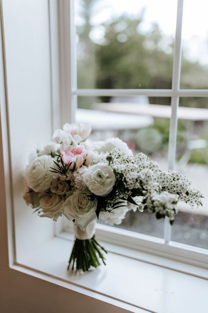 Elegant spring bridal bouquet with white ranunculus, blush tulips, and textured greenery arranged by Savannah’s Garden Floral Boutique, placed on a windowsill at Renault Winery.