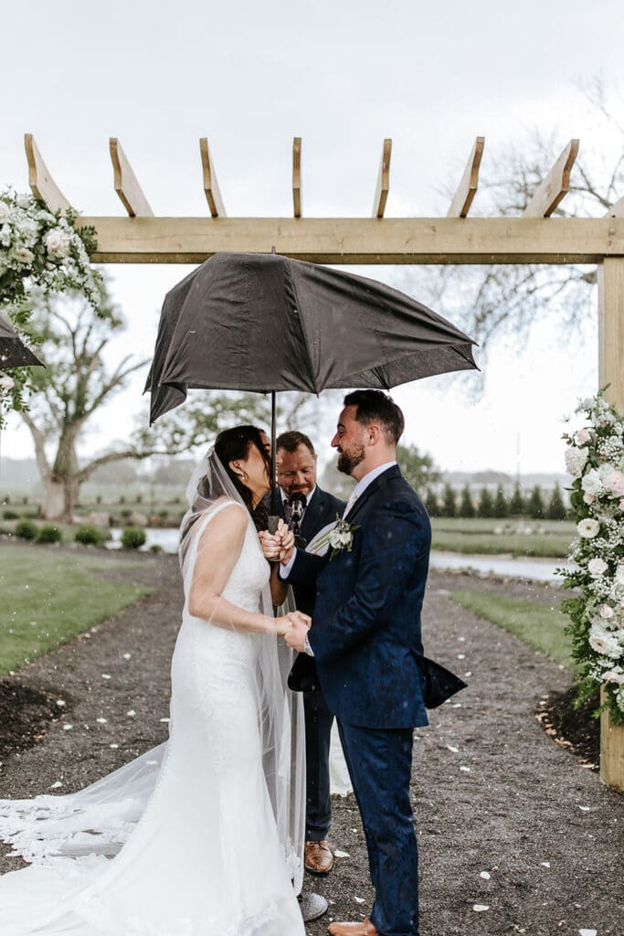 Bride and groom holding hands under an umbrella during a rainy outdoor ceremony at Renault Winery in South Jersey, with their officiant and floral arch behind them.