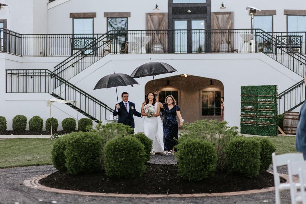 Bride and groom standing under umbrellas during a rainy outdoor ceremony at Renault Winery in South Jersey, surrounded by their bridal party and floral arbor.