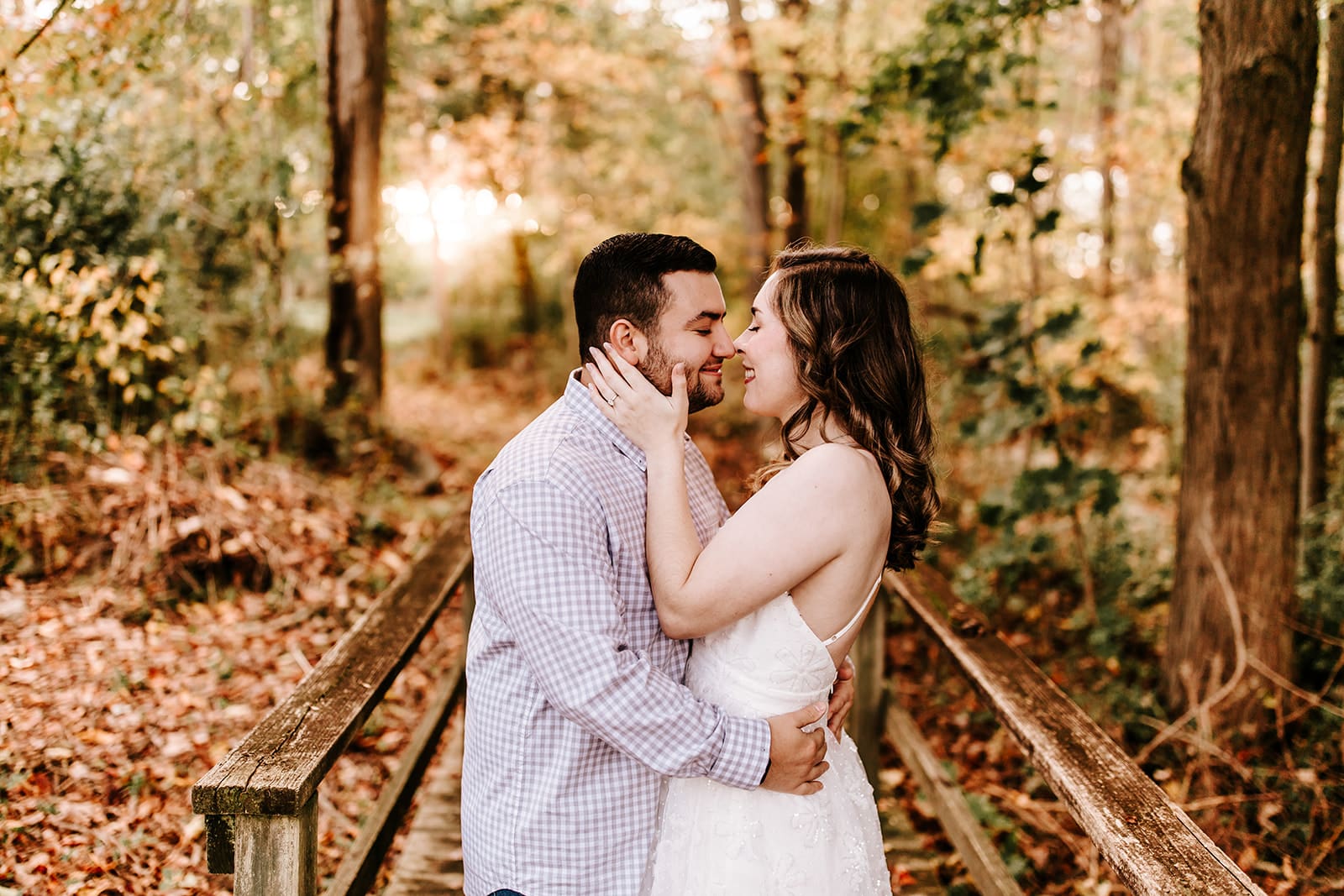 Romantic engagement photo at Springton Manor Farm in Chester County, PA. A couple shares an intimate moment on a wooden bridge surrounded by fall leaves and golden sunlight. The woman wears a white floral dress while gently holding her partner’s face. A cozy, emotional scene in a peaceful woodland setting.