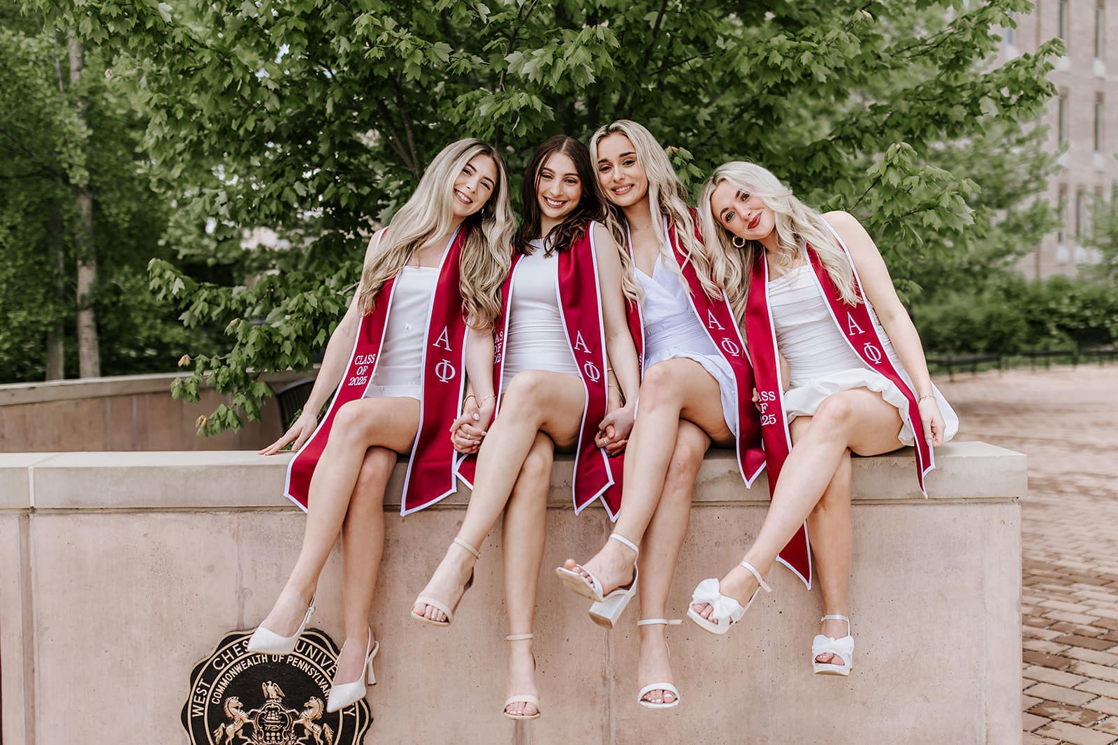Four West Chester University graduates wearing white dresses and maroon Alpha Phi stoles, sitting on a stone ledge holding hands in front of the WCU seal during spring graduation photos.
