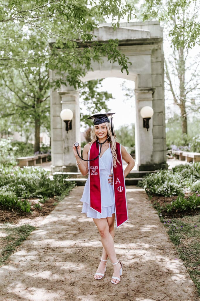 West Chester University nursing graduate holding a stethoscope in front of the historic senior arch walkway, wearing a maroon Alpha Phi stole.