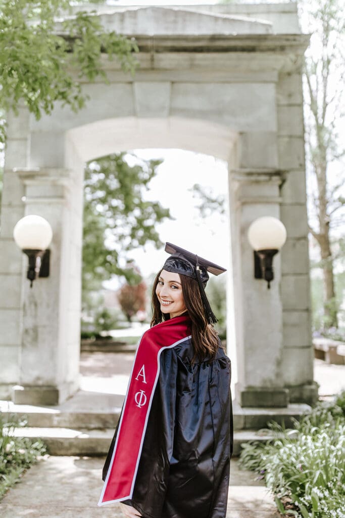 A West Chester University graduate in cap and gown smiling over her shoulder while walking through the iconic senior arch near Asplundh Concert Hall.