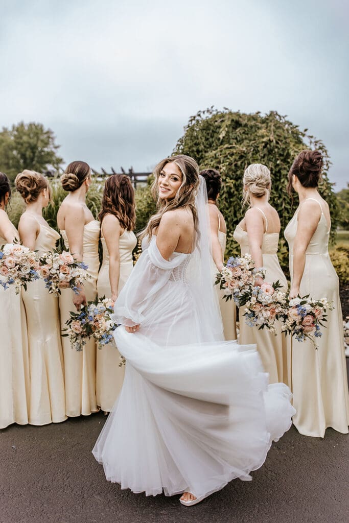 Bride twirling in an off-the-shoulder gown in front of her bridesmaids dressed in champagne satin, holding pastel floral bouquets at Bogey’s Lakeside in Pitman, NJ.