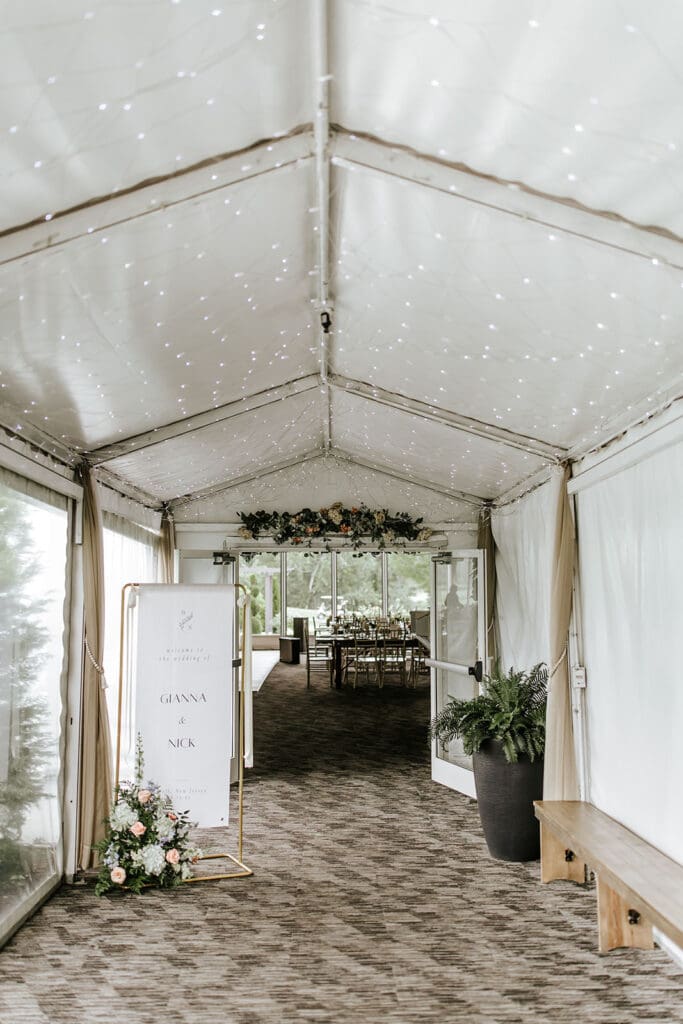 Wedding welcome sign for Gianna and Nick displayed at the entrance of a twinkle-lit reception tent at Bogey’s in Pitman, NJ, with elegant florals and candlelit dinner tables visible through the glass doors.