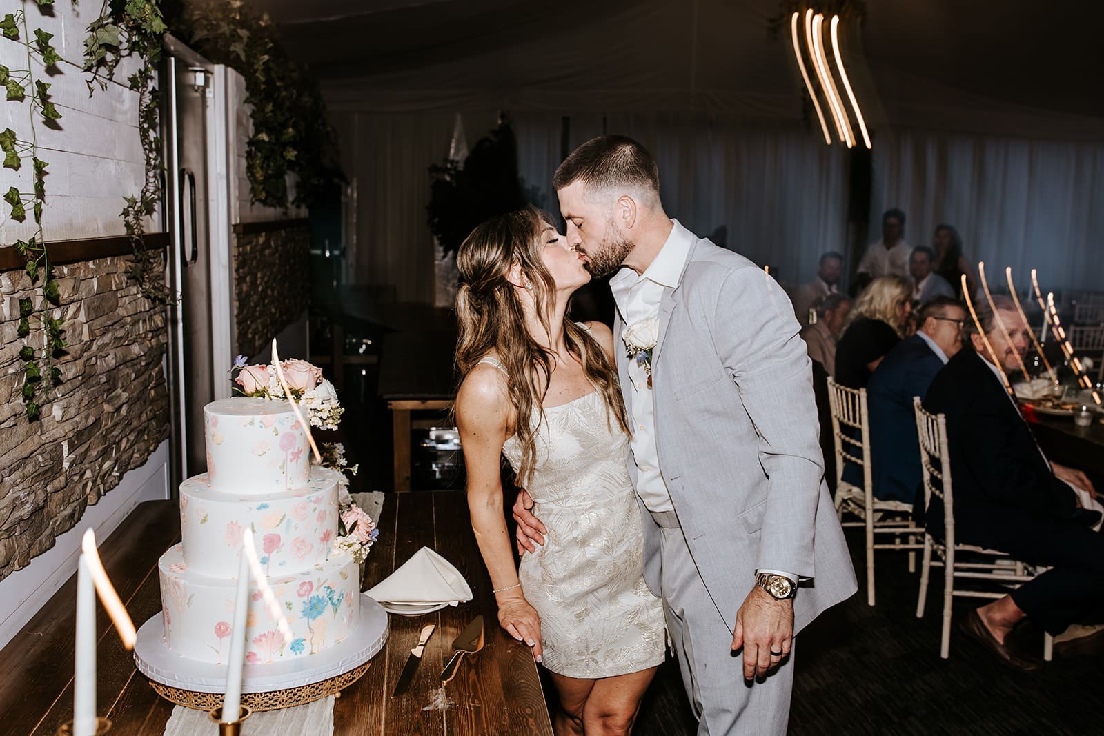 Bride and groom share a kiss next to their hand-painted floral wedding cake during the reception at Bogey’s in Pitman, NJ. The bride wears a chic mini dress while guests enjoy the celebration in the background.