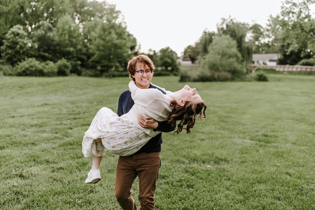 Couple laughing as the groom-to-be playfully lifts his fiancée in a wide green field at Historic Poole Forge in Chester County, PA during their spring engagement session.