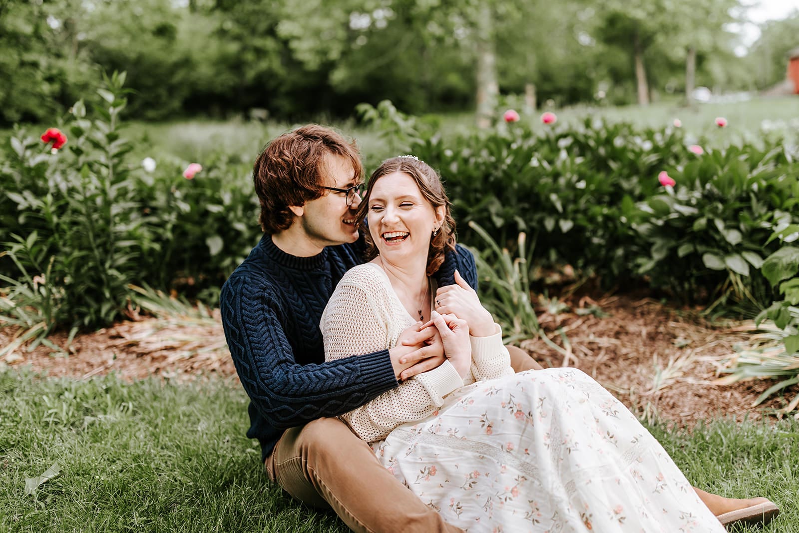 Engaged couple sitting closely and laughing together in the peony garden at Historic Poole Forge, one of Chester County’s hidden gem locations for romantic engagement photography.