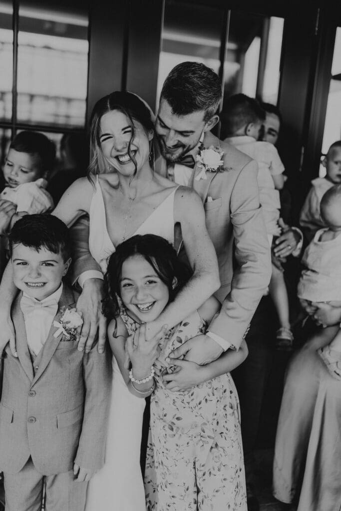 Candid black and white photo of the bride and groom hugging their children and smiling with extended family gathered around during their Westwynd Gardens wedding.